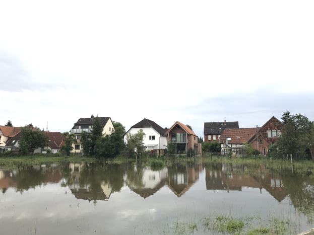 Flutstraße mit teilweise unter Wasser stehenden Häusern, umgeben von Vegetation und Strommasten, unter einem sichtbaren Himmel.