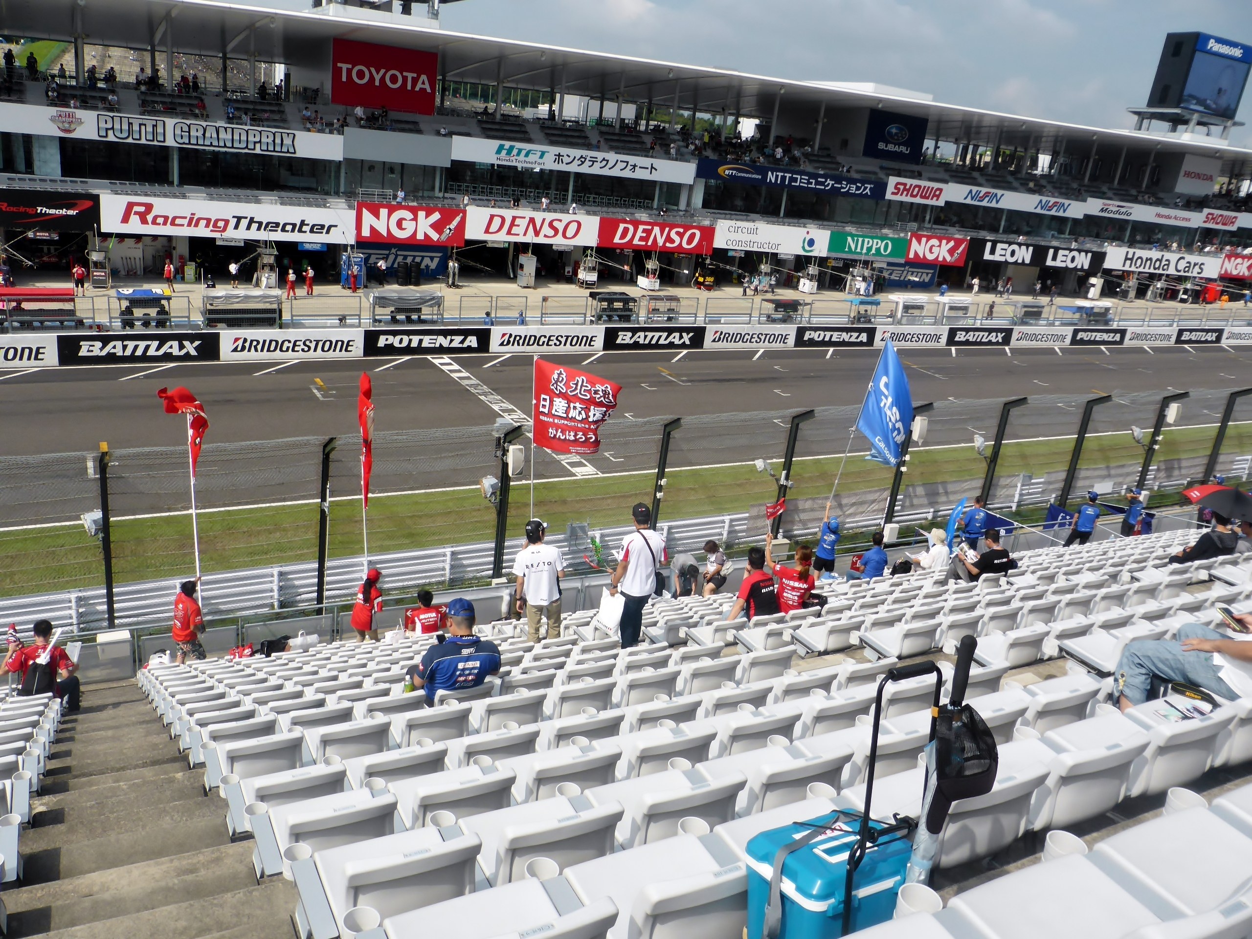 Eine Gruppe von Menschen sitzt auf Tribünen bei einer Rennstrecke mit Fahnen, Texttafeln, Geländern und Stufen, mit einem Stadion und einem bewölkten Himmel im Hintergrund.