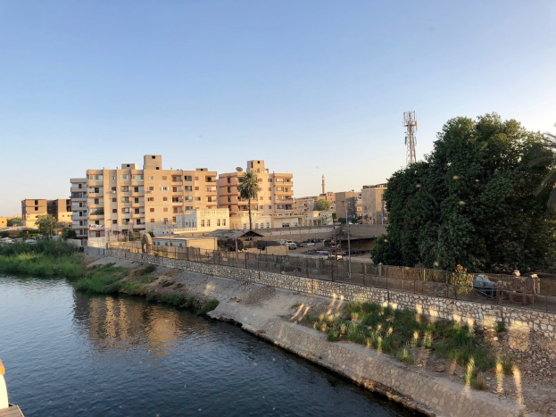 Fluss durch eine Stadt mit hohen Gebäuden, einer Brücke auf der linken Seite, umgeben von Grünflächen, Fahrzeugen auf der Straße und Türmen im Hintergrund unter einem klaren blauen Himmel.