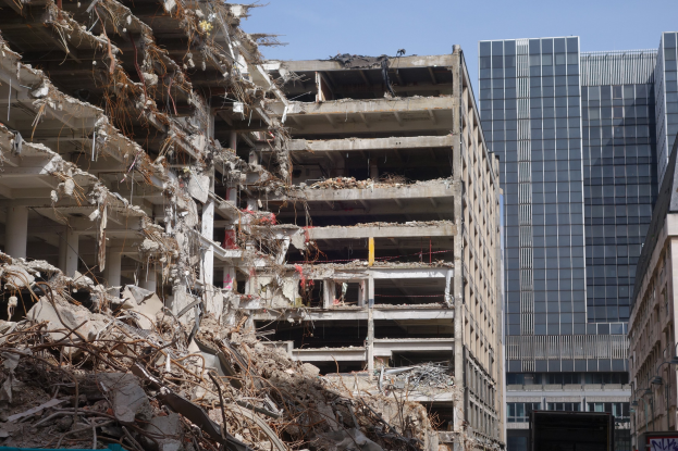 Ein großes abgerissener Gebäude in einer städtischen Umgebung umgeben von intakten Strukturen, Straßeninfrastruktur, Fahrzeugen, Bäumen und einem klaren Himmel.