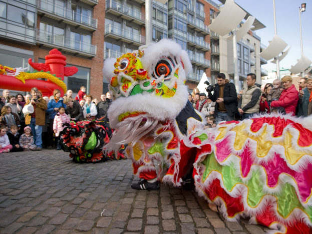 Ein lebendiges chinesisches Neujahrsfest in Amsterdam mit einer Löwen-Tanzvorstellung vor einer Zuschauermenge, darunter einige mit Kameras, vor einer Kulisse aus Gebäuden, Laternenmasten und einem klaren blauen Himmel.