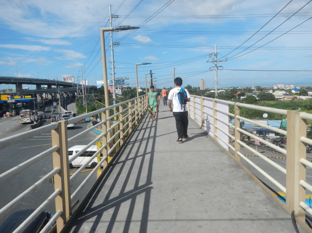 Menschen auf einer Fußgängerbrücke über einer vielbefahrenen Straße mit Fahrzeugen darunter, flankiert von Geländern und Strommasten mit Drähten, vor dem Hintergrund von Bäumen, Gebäuden und einem bewölkten Himmel.