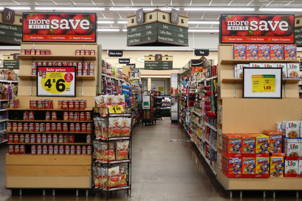 Ein Kiosk-Innenraum mit verschiedenen Lebensmitteln auf Regalen, Regalen und dem Boden, Deckenleuchten und Informationsschildern, mit einer Person im Hintergrund.