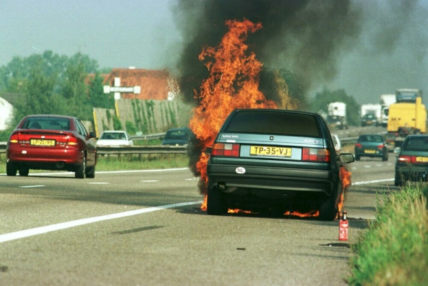 Ein Auto in Flammen auf dem Seitenstreifen umgeben von anderen Fahrzeugen mit Bäumen, Gebäuden und einem klaren blauen Himmel im Hintergrund und einem Feuerlöscher auf der rechten Seite.