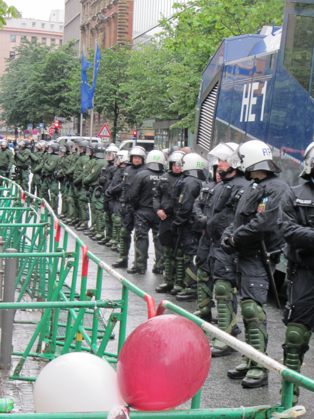 Eine Gruppe von Polizisten in Helmen und mit Waffen vor einer Barriere auf einer Straße mit Geländern und Luftballons, mit Bäumen, Gebäuden, Flaggen und einem klaren blauen Himmel im Hintergrund.