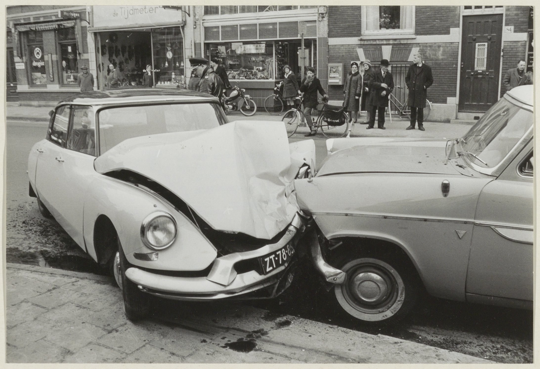 Schwarz-weiß-Foto von zwei beschädigten Autos am Straßenrand mit Menschen und Fahrrädern in der Nähe und Gebäuden im Hintergrund.