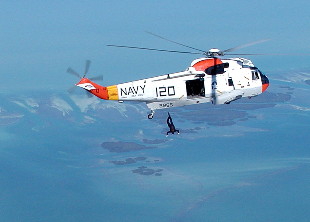 Ein Rettungshubschrauber fliegt über Wasser mit einer Person darunter, vor einem Himmel-Hintergrund.