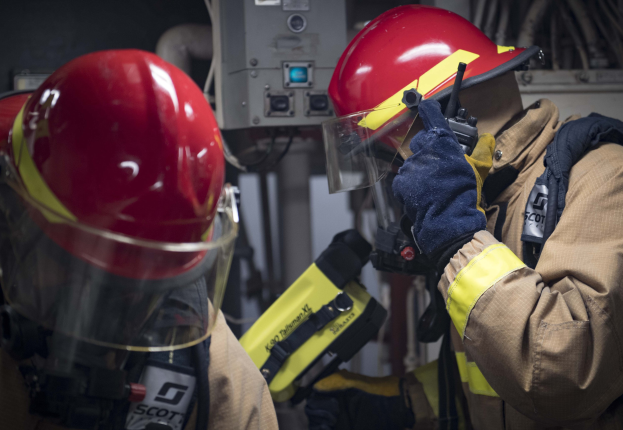 Zwei Feuerwehrleute in Schutzausrüstung arbeiten an einem Hydranten während einer Trainingsübung mit Maschinen und Kabeln im Hintergrund.