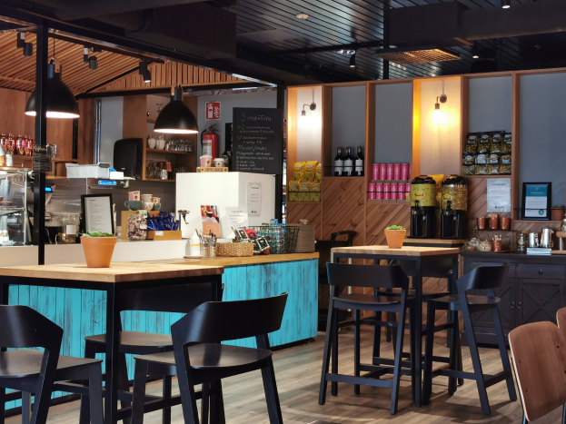 Innenraum eines Restaurants mit Holztischen, Stühlen, einer Topfpflanze auf einem Tisch, einer Tafel, einem Kühlschrank, einem Feuerlöscher, Regalen mit Flaschen, Schränken mit Gegenständen, einem Foto auf der Wand, Deckenleuchten und Lampen.