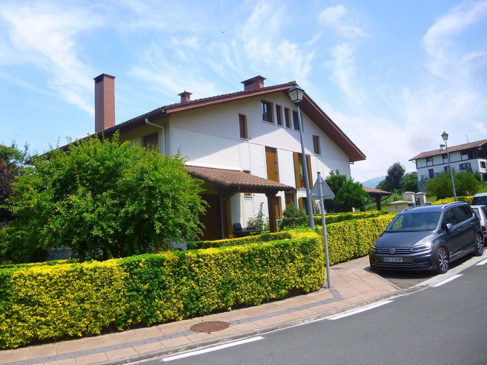 Ein zweistöckiges Villa zum Verkauf in Lago di Garda, Italien, mit einem Auto auf der Straße davor geparkt, umgeben von Grün und Laternen, unter einem teilweise bewölkten Himmel.