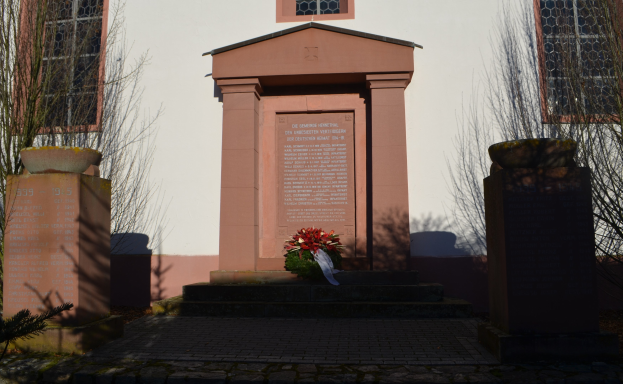 Ein Gebäude mit Fenstern, ein Denkmal mit Text, ein Blumenstrauß auf dem Boden, Topfpflanzen, Bäume, ein bewölkter Himmel und ein Grab mit einem Kranz davor.