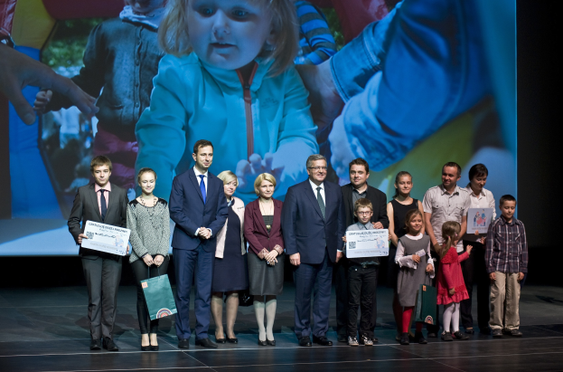 Gruppe von Menschen auf einer Bühne mit Taschen und Urkunden, mit einem Hintergrundbild eines Kindes und anderer Personen.