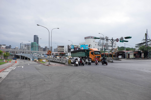 Eine belebte Stadtstraße mit Fahrzeugen wie einem Lastwagen und einem Auto, Laternenmasten, Verkehrsampeln, Schildern, einer Brücke mit Geländern, Bäumen, Gebäuden mit Fenstern und einem bewölkten Himmel im Hintergrund.