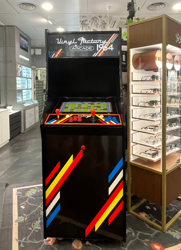 Ein Geschäft mit einem zentralen Retro-Arcade-Automaten, einer Glasvitrine auf der rechten Seite und Retro-Beleuchtung mit Schränken, einem Fernseher und anderen Gegenständen im Hintergrund.