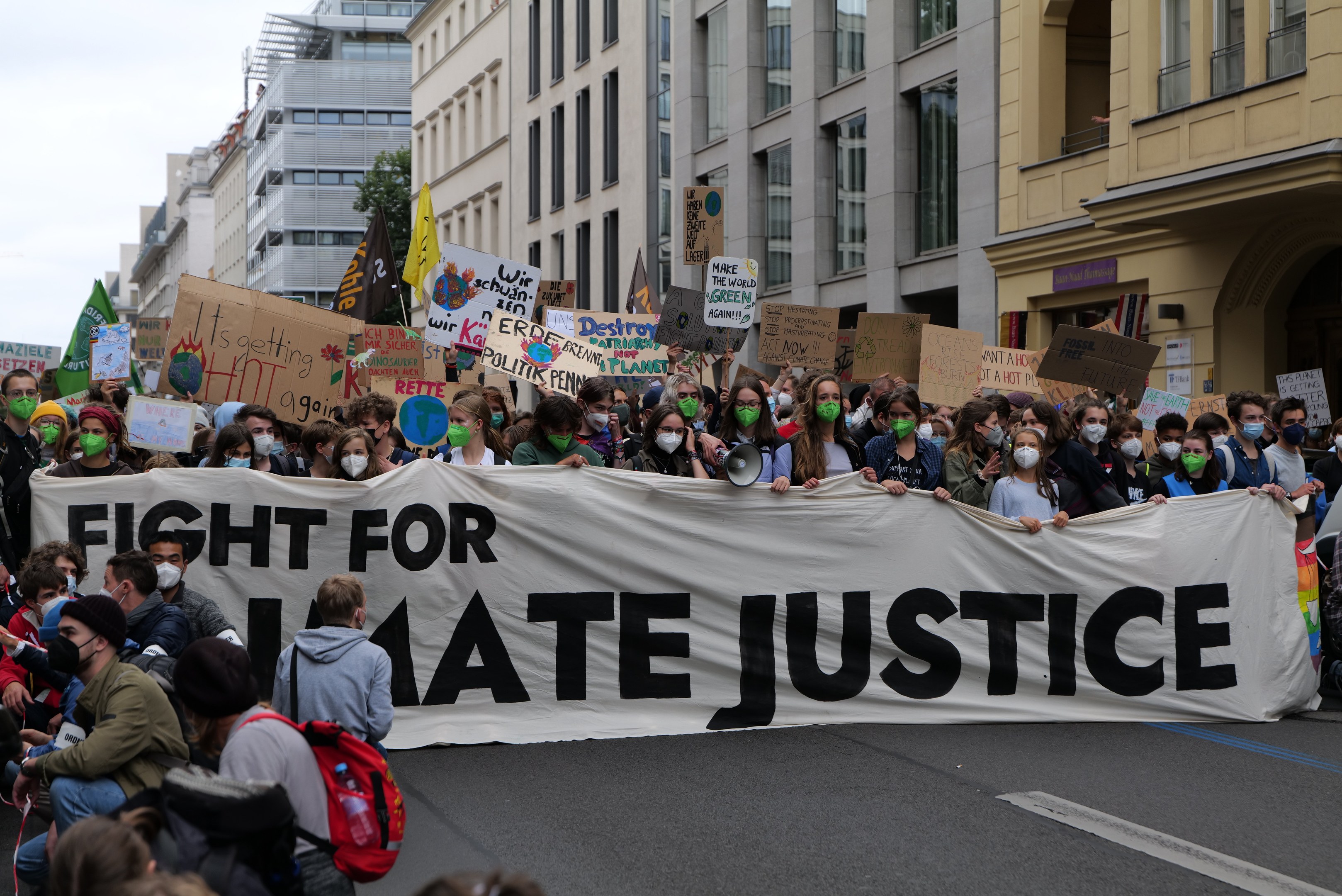 Eine große Gruppe von Menschen marschiert mit einem Banner, auf dem 'Kampf für Hassjustiz' steht, durch die Straße. Einige tragen Masken und halten Schilder oder Taschen, vor einem klaren blauen Himmel mit Gebäuden und Bäumen im Hintergrund.