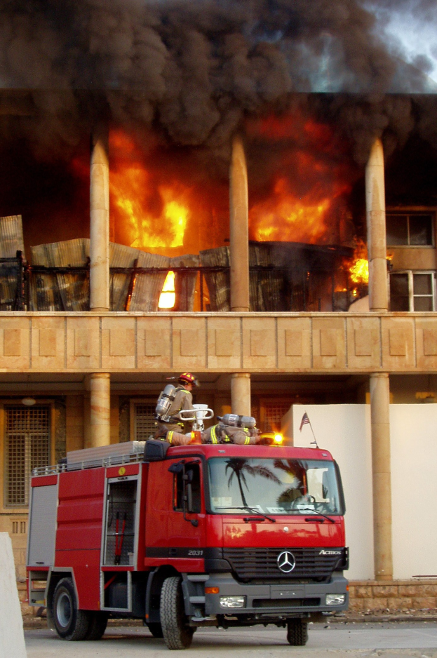 Feuerwehrfahrzeug auf der Stra├če vor einem in Flammen stehenden Geb├Ąude geparkt, mit einer Wand links.
