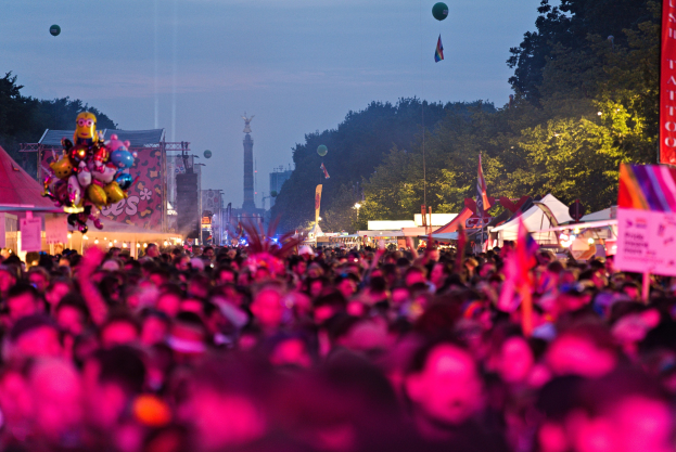 Eine große Menschenmenge bei einem nächtlichen Musikfestival, beleuchtet von bunten Lichtern und Ballons, mit Zelten, Banner, Flaggen, Bäumen, einem Turm und einem sternenklaren Himmel im Hintergrund.