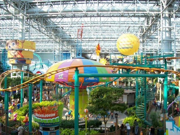 Indoor-Wasserpark mit einer Achterbahn, umgeben von Menschen, Bäumen, Pflanzen und Deckenbeleuchtung mit Stangen.