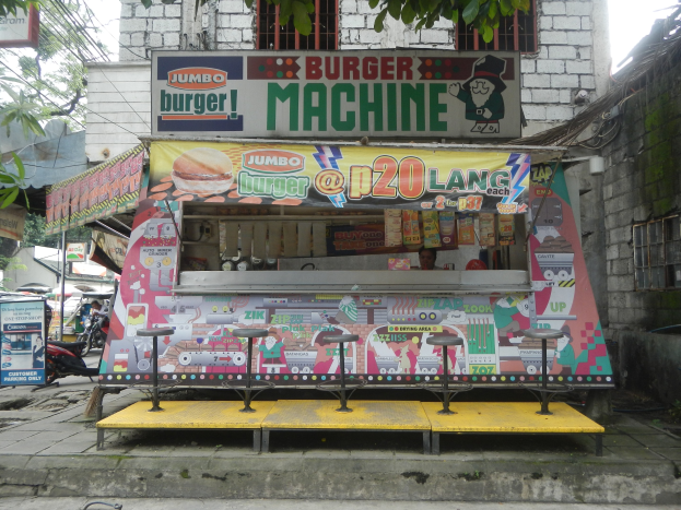 Ein leuchtend gelber und roter Foodtruck in Form eines Jumbo-Burgers, der auf einer von Gebäuden, Bäumen und Menschen umgebenen Straße geparkt ist, mit einem sichtbaren Himmel und einer Straße im Hintergrund.