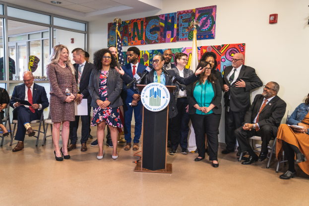 Eine Gruppe von Menschen steht und sitzt in einem Raum mit einem Podium, einem Mikrofon und einer Tafel mit der Aufschrift "Sancho-Stadtrat tritt dem Stadtrat bei", mit Flaggen, einem Feuerlöscher und Deckenleuchten im Hintergrund.