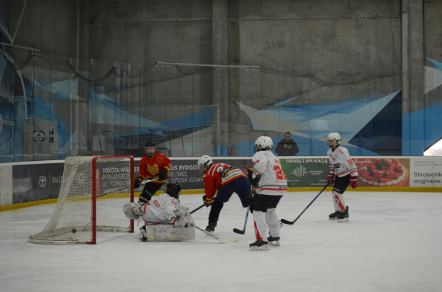 Eine Gruppe von Menschen, die Eisockey auf einem Eisplatz spielen, mit Helmen und Eishockeyschlägern, sowie einem Torpfosten auf der linken Seite und Bannern im Hintergrund.