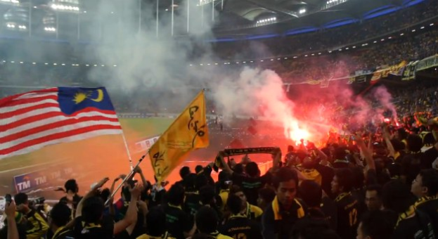 Eine große Menge Menschen in einem Stadion mit brennenden Fackeln in der Luft, einige halten Fahnen und eine Person hält eine Kamera, mit Rauch im Hintergrund und Tafeln mit der Aufschrift "Malaysia vs Malaysia Live-Streaming".