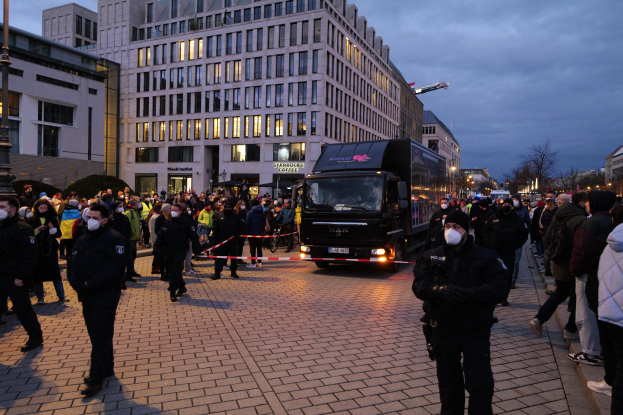 Eine Gruppe von Menschen steht in der Nähe eines Lastwagens auf einer Straße, die von Gebäuden, Laternen und Bäumen gesäumt ist, unter einem bewölkten Himmel, wobei einige Mützen und Masken tragen und ein Band auf einem Pfahl im Vordergrund zu sehen ist.
