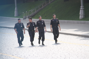 Drei Polizisten in Uniform gehen über eine Straße mit Mästängen, Geländern, Treppen und Gras im Hintergrund.