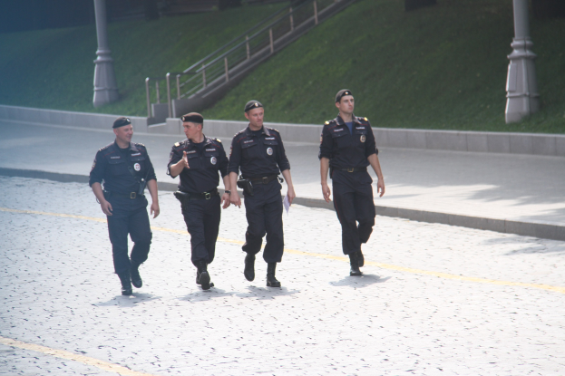 Drei Polizisten in Uniform gehen über eine Straße mit Mästängen, Geländern, Treppen und Gras im Hintergrund.