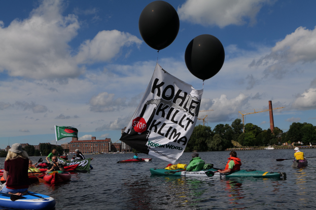 Gruppe von Menschen in Kajaks auf dem Wasser mit einem Banner, das "Kohle Kill Klima" und Paddel in den Händen hält, umgeben von Bäumen, Gebäuden, Kränen und einem klaren blauen Himmel.