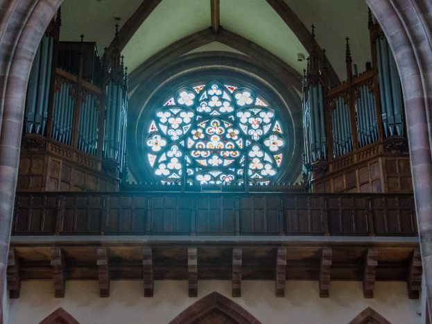 Innensicht einer Kirche mit einem zentralen Buntglasfenster, das von Holzgeländern und Säulen umgeben ist, mit verstreuten Gegenständen und einer Wand unten.