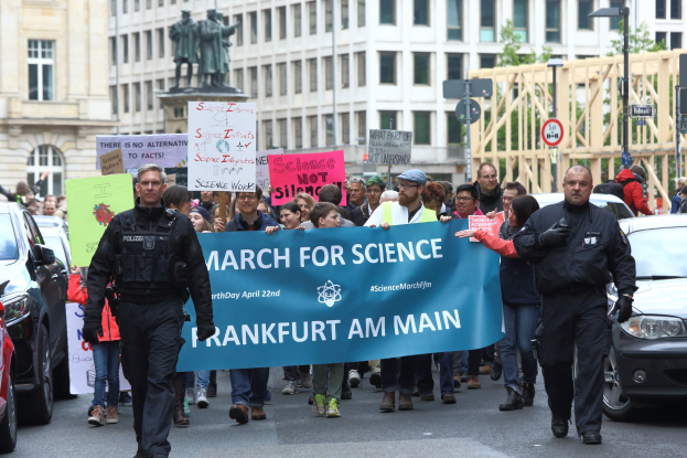 Gruppe von Menschen marschiert auf der Straße mit einer "March for Science Frankfurt am Main"-Plakette, Autos, Gebäude, Statuen, Laternen, Schilder und Bäume im Hintergrund.
