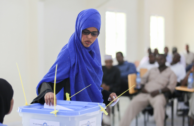 Eine Frau in einem blauen Kopftuch gibt ihre Stimme an einer Wahllokal ab, während andere in der Nähe sitzen.