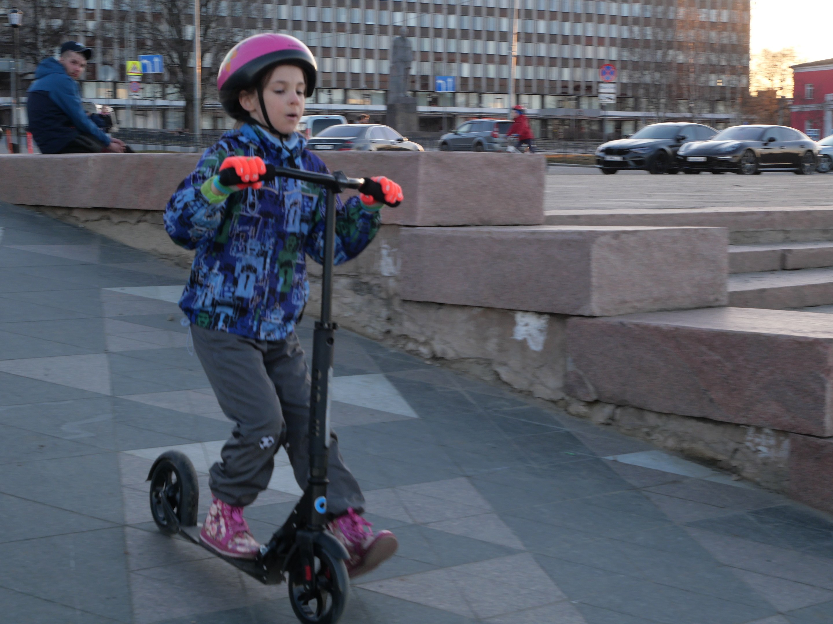 Ein junger Junge in einem Helm und Handschuhen fährt auf einem Gehweg mit Stufen, Fahrzeugen, Menschen, Bäumen, Pfosten, Brettern, Gebäuden und einem klaren blauen Himmel im Hintergrund.