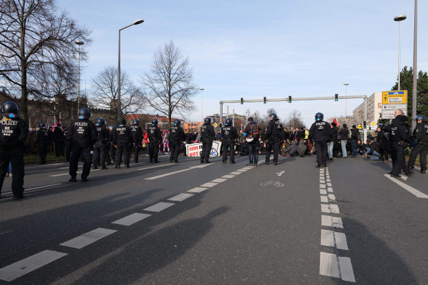 Gruppe von Polizisten in schwarzer Uniform und Helmen auf der Seite einer Straße mit Laternenmasten, Ampeln, Bäumen, Gebäuden und einem klaren blauen Himmel im Hintergrund.