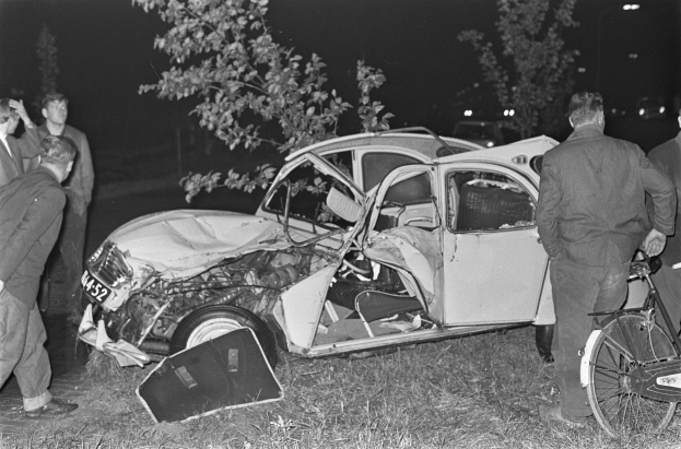Schwarzes und weißes Bild eines auf der Straße liegengebliebenen Autos mit mehreren Menschen in der Nähe, einer hält ein Fahrrad, und beleuchtete Bäume im Hintergrund.