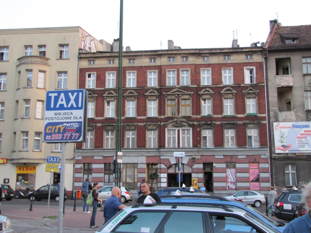 Eine belebte Stadtstraße mit einem geparkten Taxi, fahrenden Fahrzeugen, Fußgängern, einem Wegweiser, Gebäuden und einem sichtbaren Himmel.