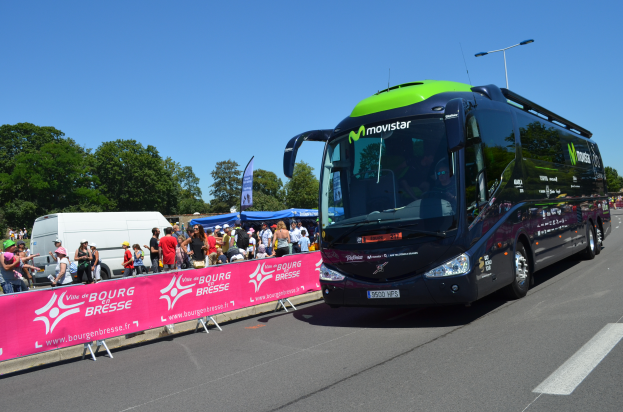 Ein schwarzer und grüner Bus fährt auf einer Straße neben einer Menschenmenge, einige tragen Mönen, mit einem Banner auf der linken Seite und Bäumen unter einem klaren blauen Himmel im Hintergrund.