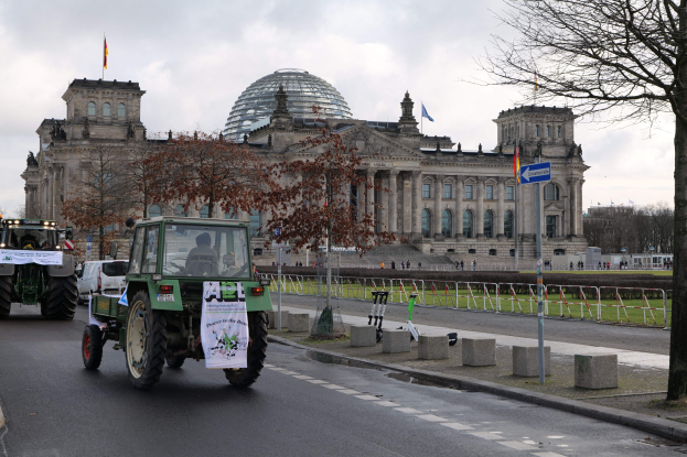 Gruppe von Traktoren fährt vor dem Reichstaggebäude in Berlin, Deutschland, die Fenster, Säulen und Flaggen des Gebäudes, umgeben von Bäumen, Geländern und anderen urbanen Elementen unter einem bewölkten Himmel.