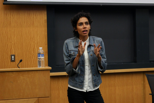 Frau am Rednerpult mit Mikrofon und Flasche, die vor einem Projektionsschirm eine Rede hält, mit einem Stuhl rechts daneben und einer hölzernen Wand im Hintergrund.