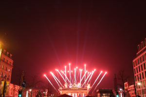 Eine nächtliche Stadtstraße mit Gebäuden, Bäumen, Laternen, Ampeln, Schildern und Menschen im Vordergrund und einem Feuerwerk am Himmel.