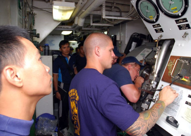 Männer in blauen T-Shirts stehen um ein Schiffskontrollpult herum, einige mit sichtbaren Tattoos an den Händen, während Meter und elektronische Geräte rechts zu sehen sind. Der Hintergrund zeigt eine Wand mit Rohren und Deckenleuchten.
