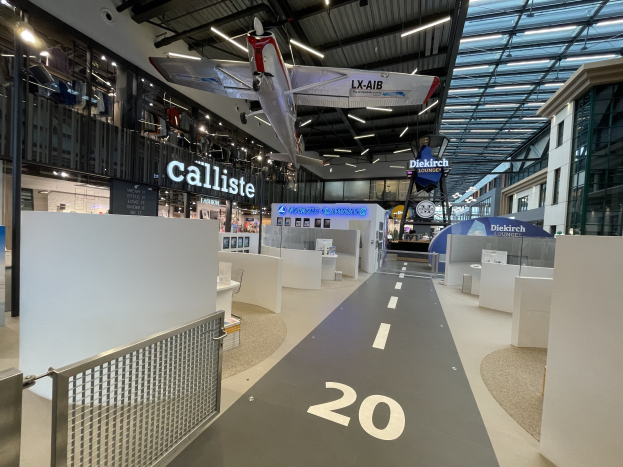 Großer Raum mit weißen Wänden, ein Flugzeug an der Decke, ein Geländer auf der linken Seite, verstreute Objekte auf der rechten Seite, Texttafeln im Hintergrund und Deckenlampen an Metallstangen.