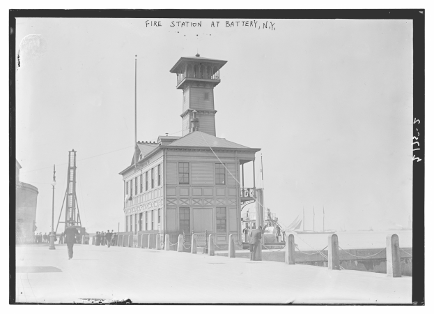 Schwarzes und weißes Foto einer Feuerwache in Battery, New York, mit einem Gebäude, Menschen, Pfählen, einem Zaun, Booten auf dem Wasser und einem bewölkten Himmel.