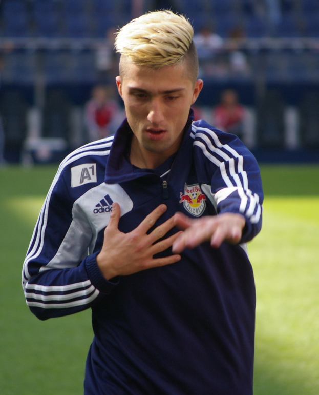 Ein junger Mann in einem blauen T-Shirt steht auf einem Fußballfeld mit ausgestreckten Armen und sieht entschlossen aus, während der Hintergrund unscharf ist.