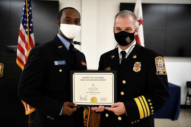 Zwei Männer in Feuerwehrausstattung stehen nebeneinander, tragen Masken und halten ein Zertifikat, im Hintergrund sind Fahnen und Möbel zu sehen; der Mann links ist der Feuerchief des Bezirks Washington und erhält eine Auszeichnung.