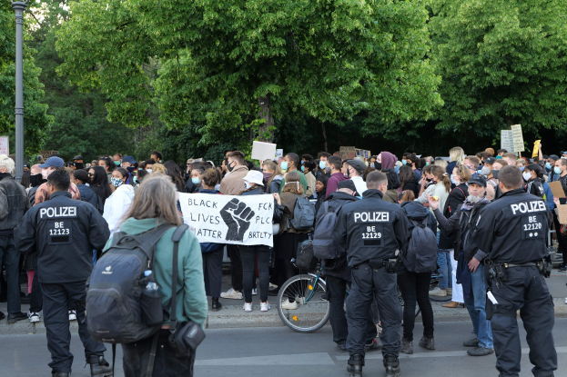Eine große Gruppe von Menschen nimmt an einer Black Lives Matter Demo in Berlin teil, einige halten Schilder und tragen Mützen und Taschen, mit einem Fahrrad im Vordergrund und Bäumen und einem Pfahl im Hintergrund.