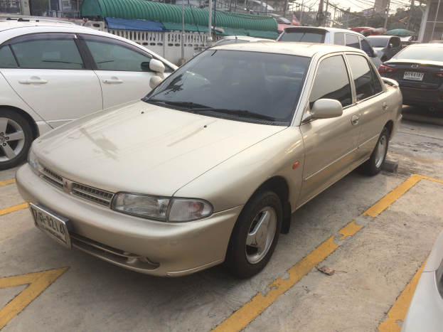 Ein goldfarbener Wagen auf einem Parkplatz mit anderen Autos, Gebäuden, Masten, Drähten, Bäumen und einem klaren blauen Himmel.