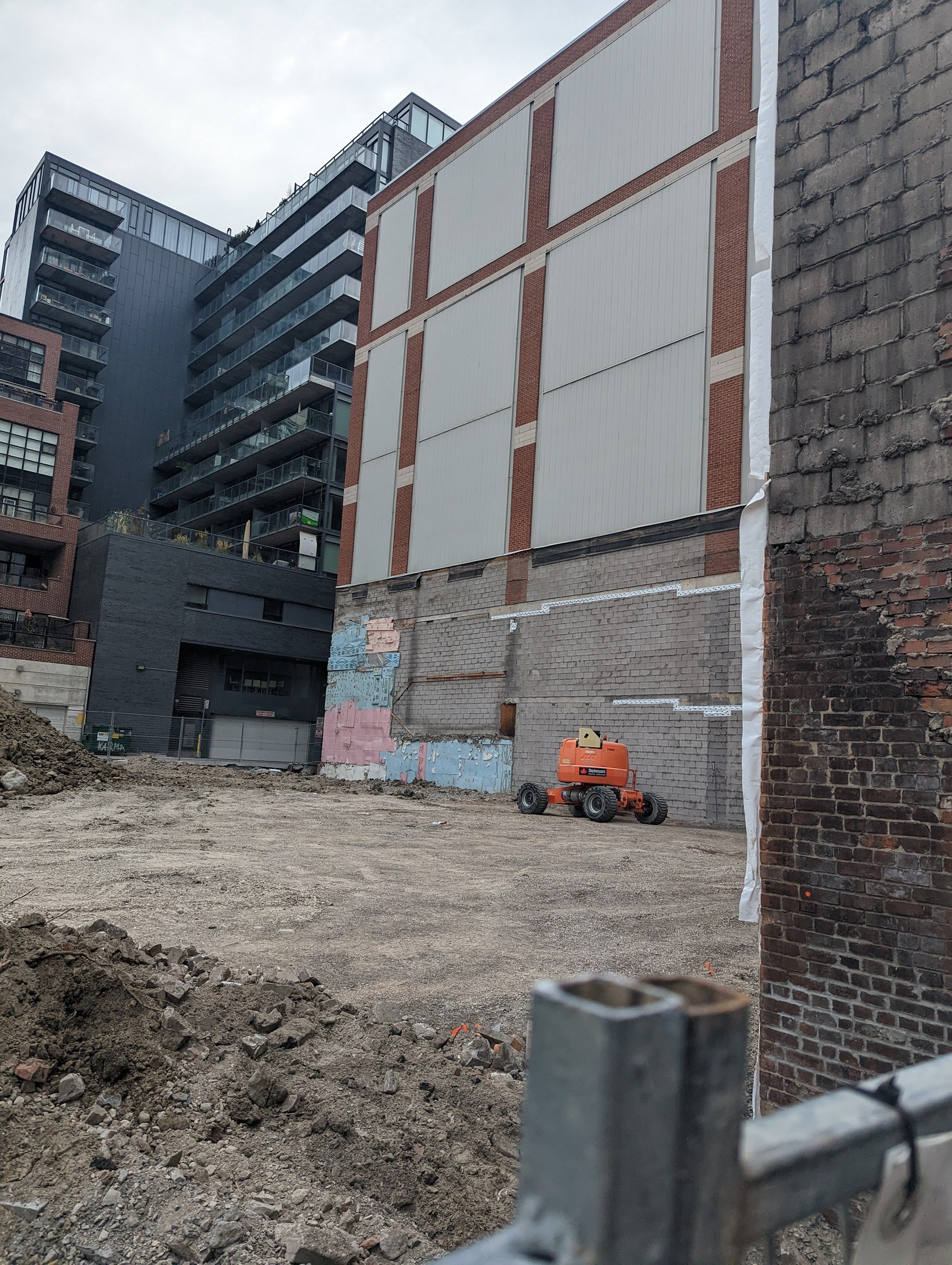 Baustelle mit einem teilweise abgerissenen Gebäude im Hintergrund, einem Fahrzeug auf dem Boden, einem Metallzaun und einem bewölkten Himmel.