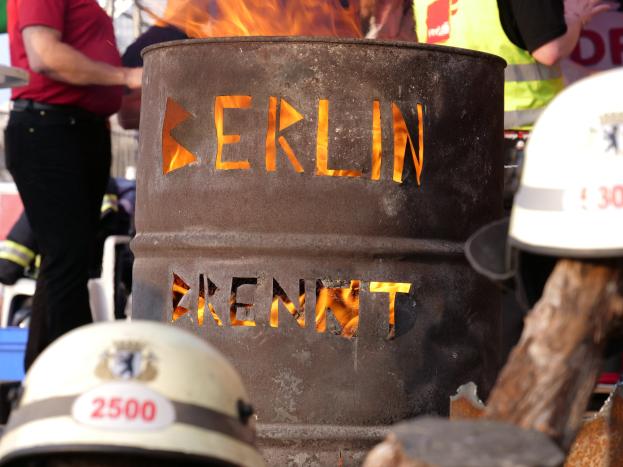 Eine Gruppe von Menschen steht in der Nähe eines Feuerhydranten mit der Aufschrift "Berlin", im Vordergrund brennt ein Fass, Helme unten und ein hölzernes Objekt rechts.
